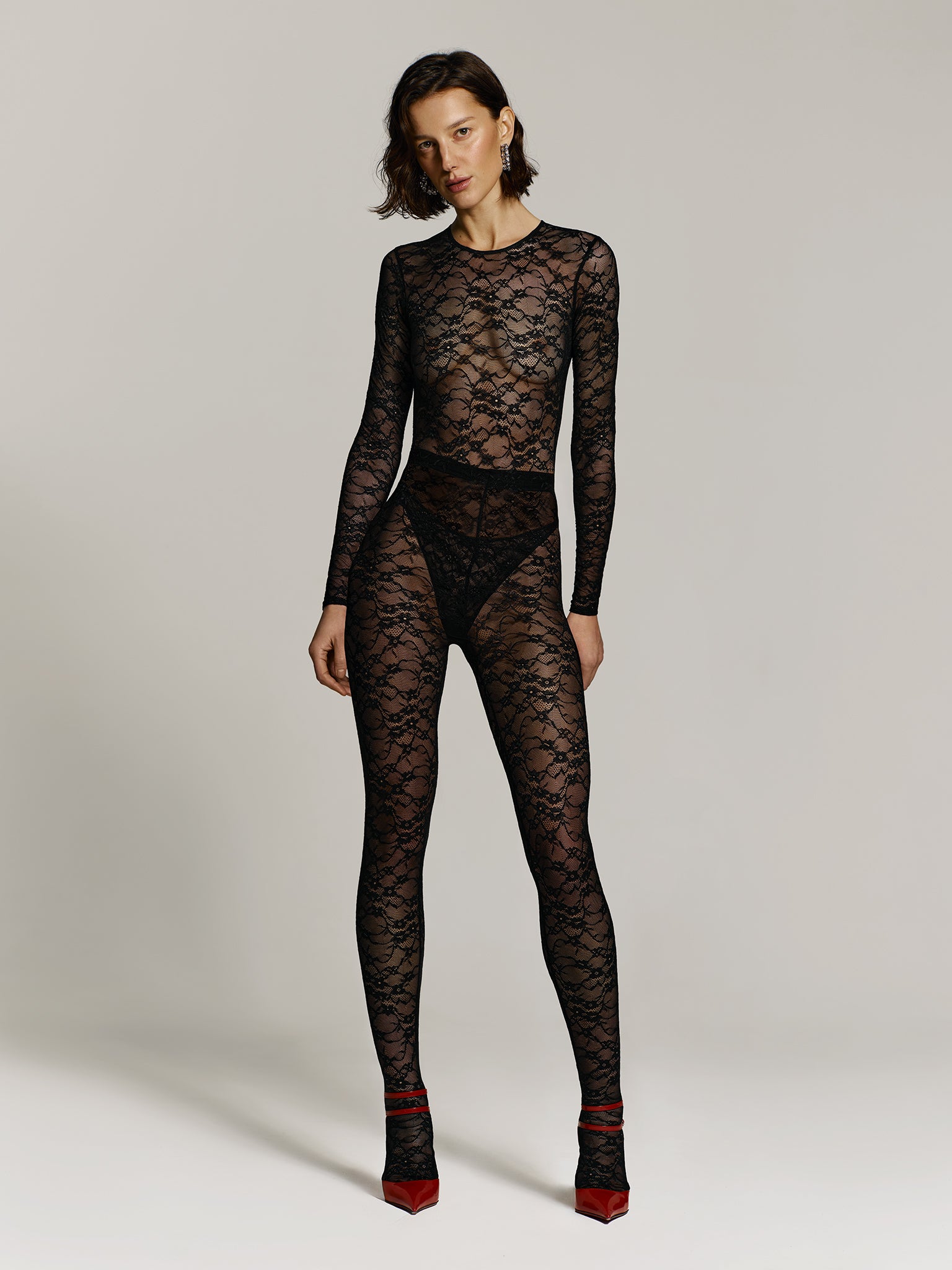 Full shot of a girl in a black lace long sleeved bodysuit, black lace high rise leggings and red high heels