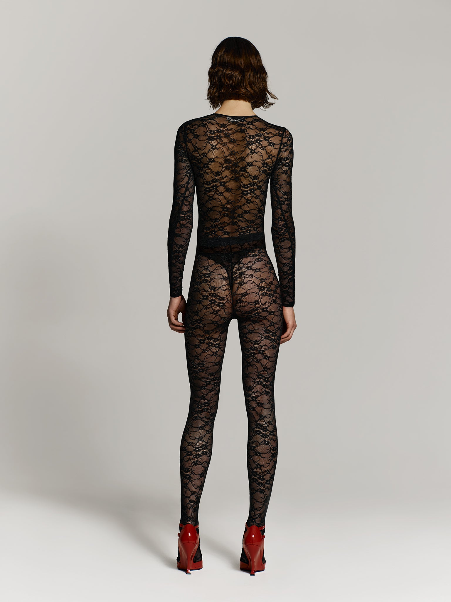Full shot of a girl facing back in a black lace long sleeved bodysuit, black lace high rise leggings and red high heels