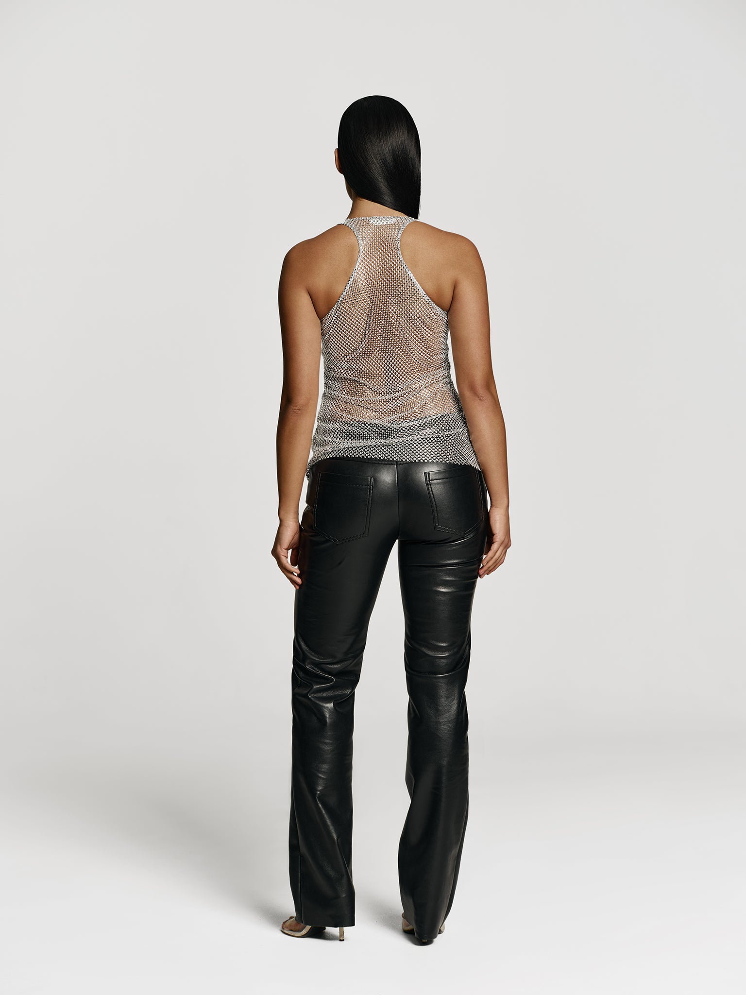Full shot of a girl facing back in a grey net tank top decorated with rhinestones and black leather medium rise straight leg pants