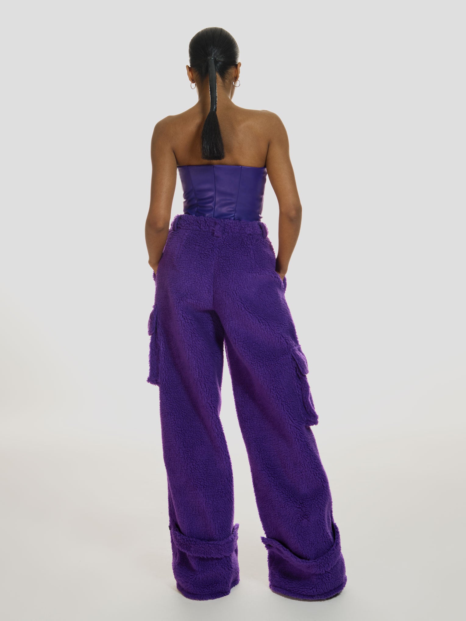 Full shot of a girl facing back with her hands in pockets wearing a purple vegan leather tube top and purple polar fleece cargo pants
