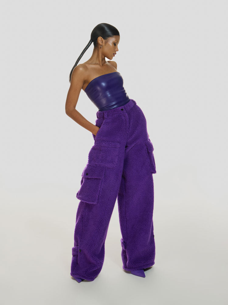 Full shot of a girl with her hands in pockets wearing a purple vegan leather tube top and purple polar fleece cargo pants