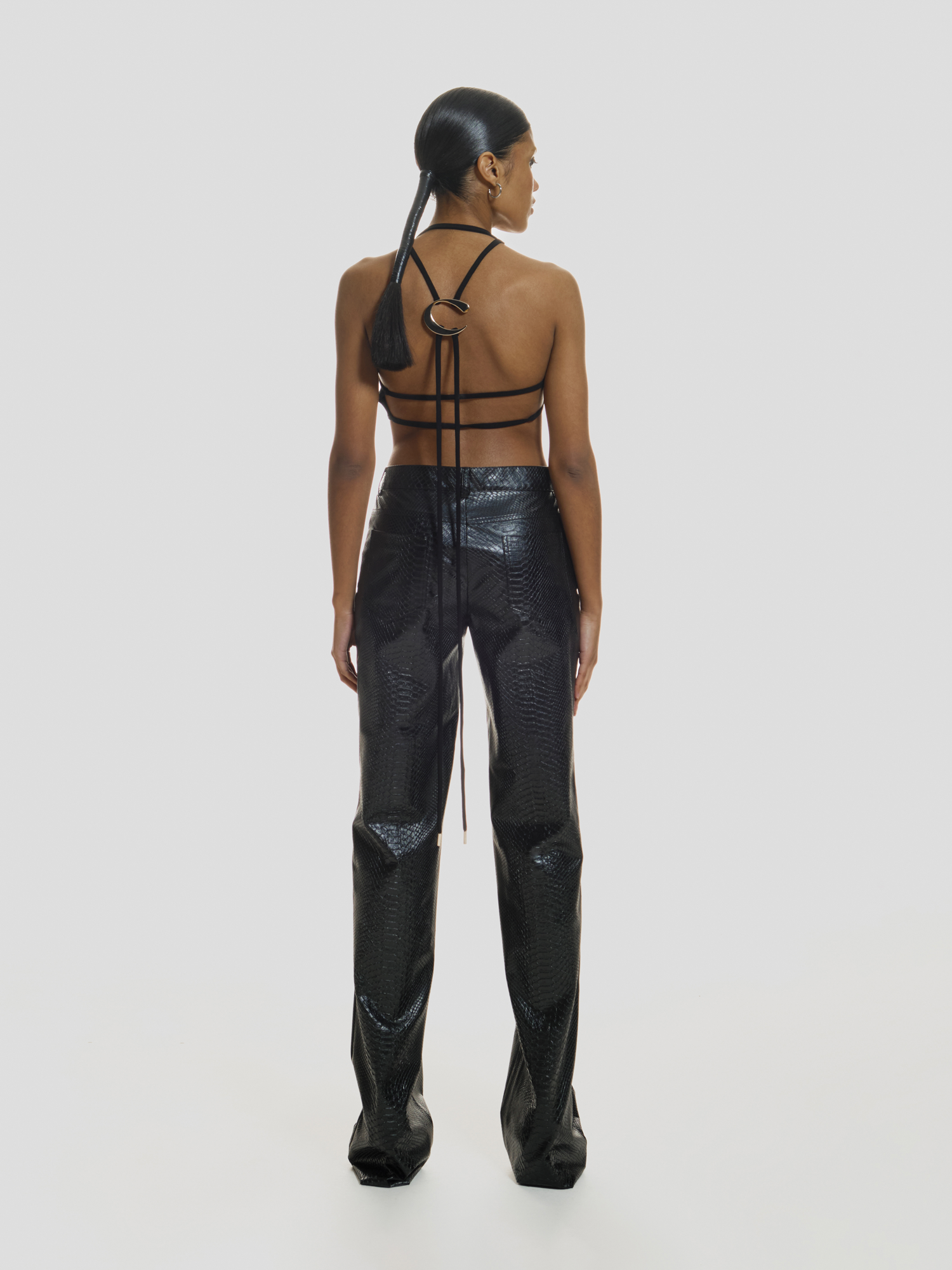 Full shot of a girl facing back in a black crop top with extended laces and black crocodile printed vegan leather pants with mid rise