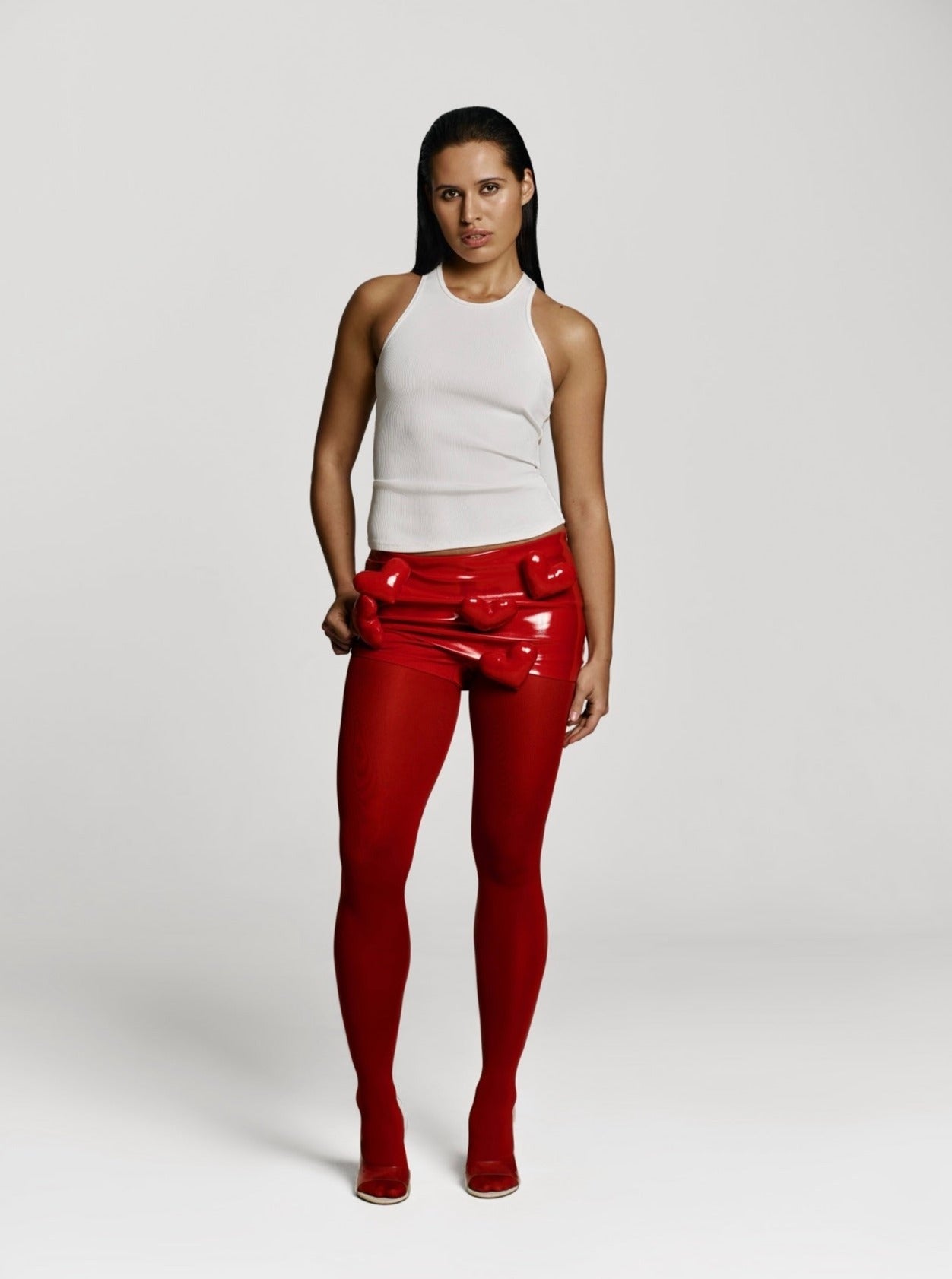 Full shot of a girl in a white viscose tank top, red patent vegan leather mini skort decorated with hearts and red tights