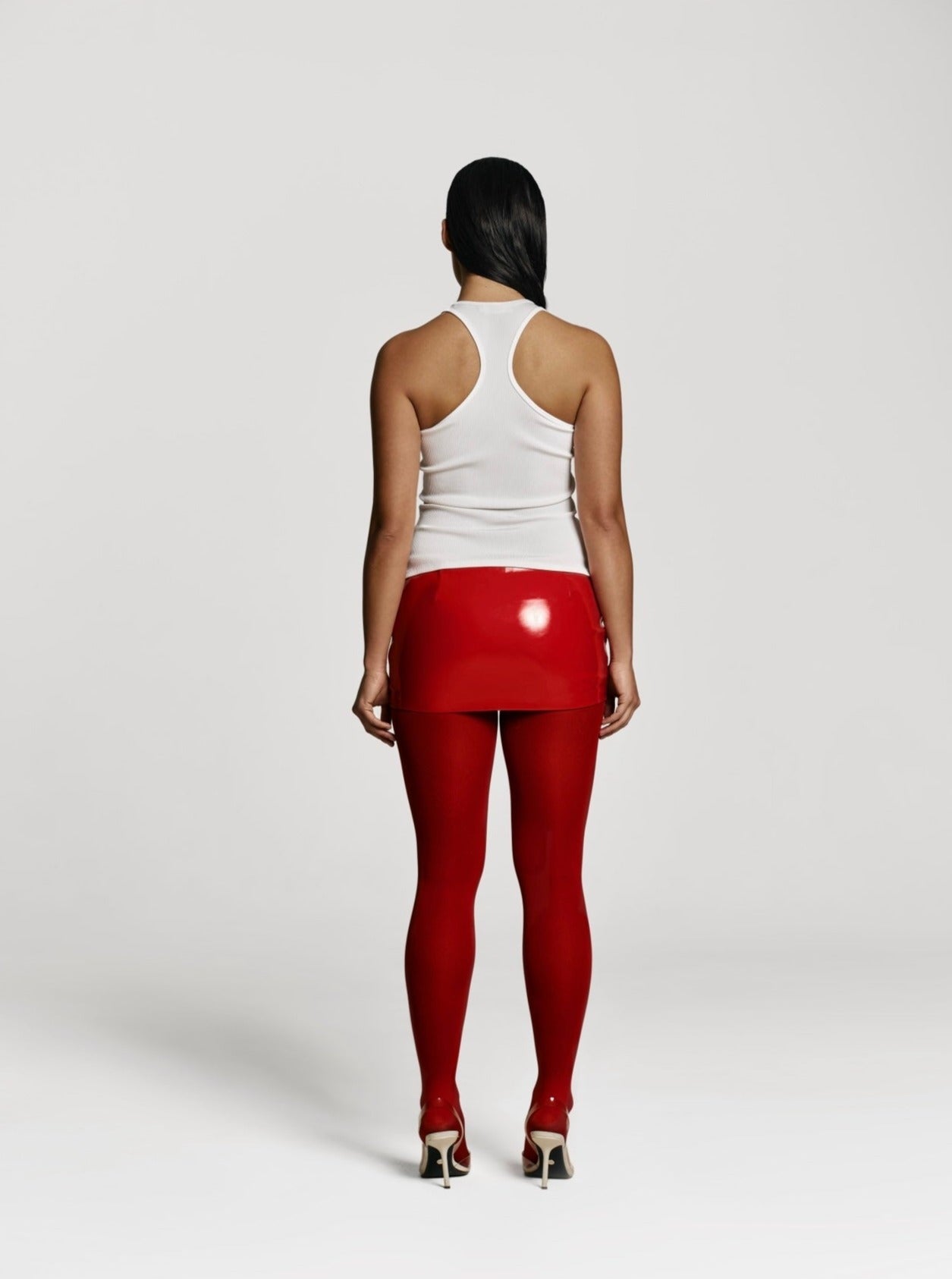 Full shot of a girl facing back in a white viscose tank top, red patent vegan leather mini skort decorated with hearts and red tights