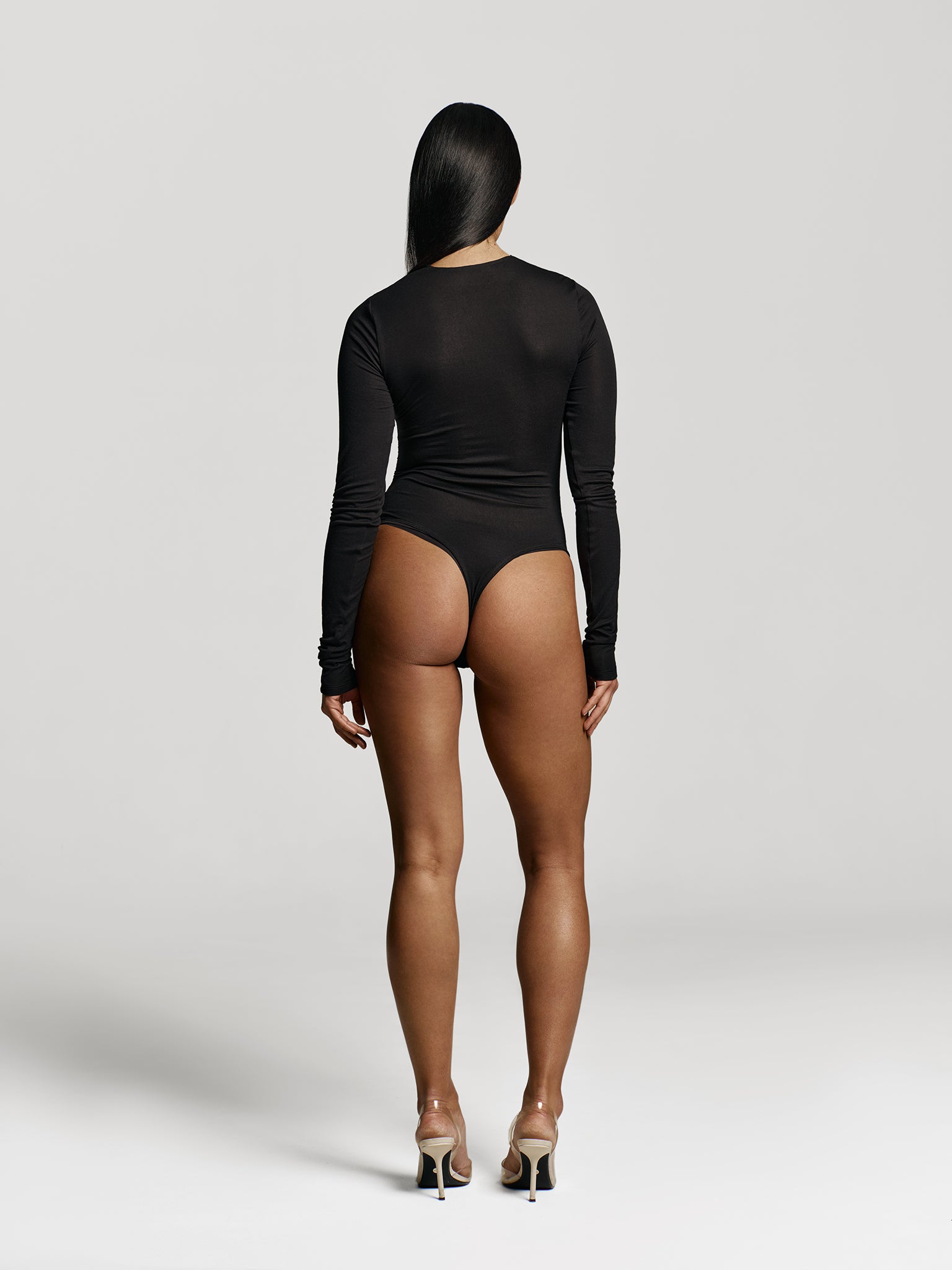 Full shot of a girl facing back in a black long sleeved bodysuit with draping at the sides and high round neckline 