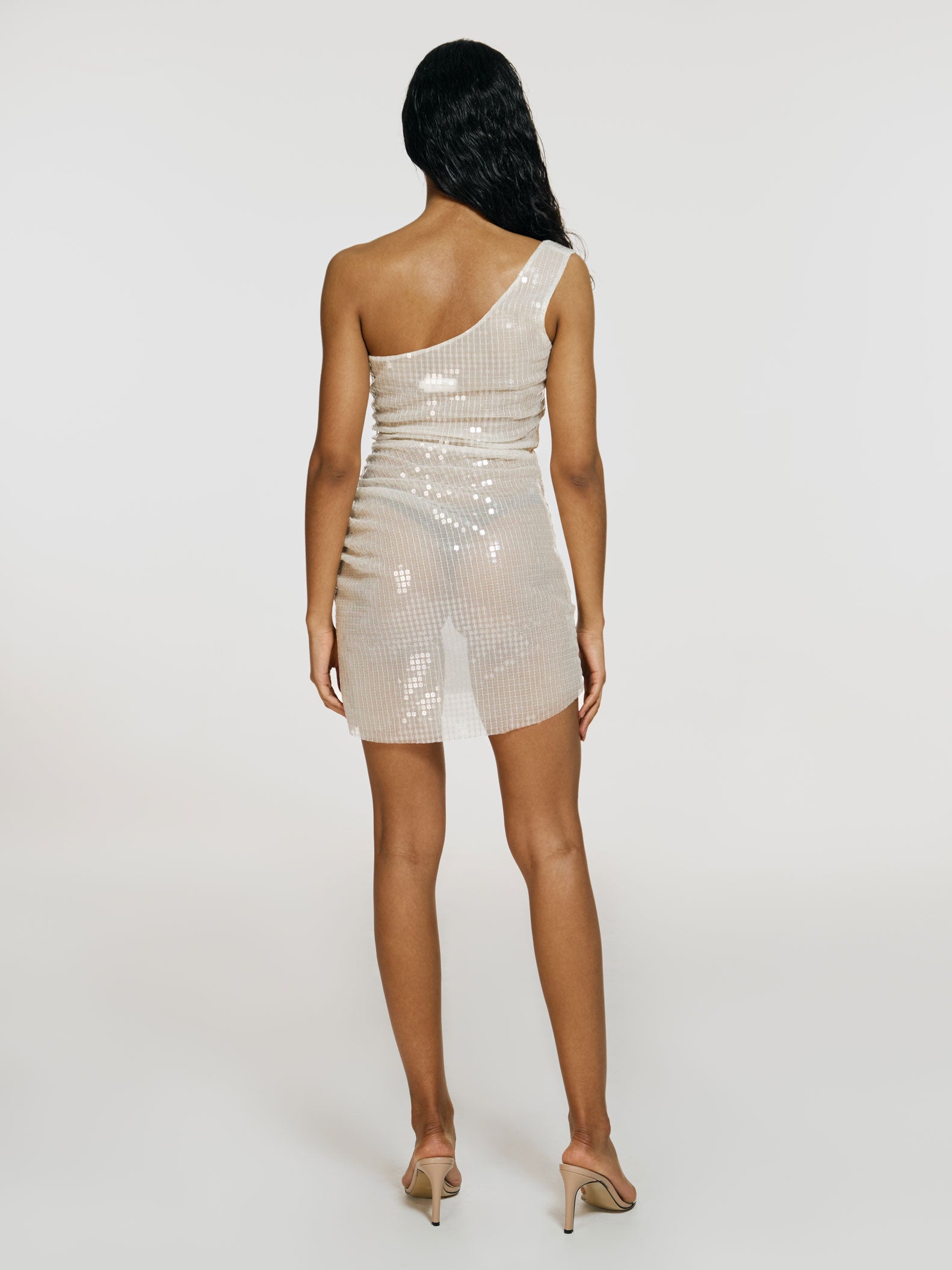 Full shot of a girl facing back in a beige semi-transparent sequinned one shoulder mini dress