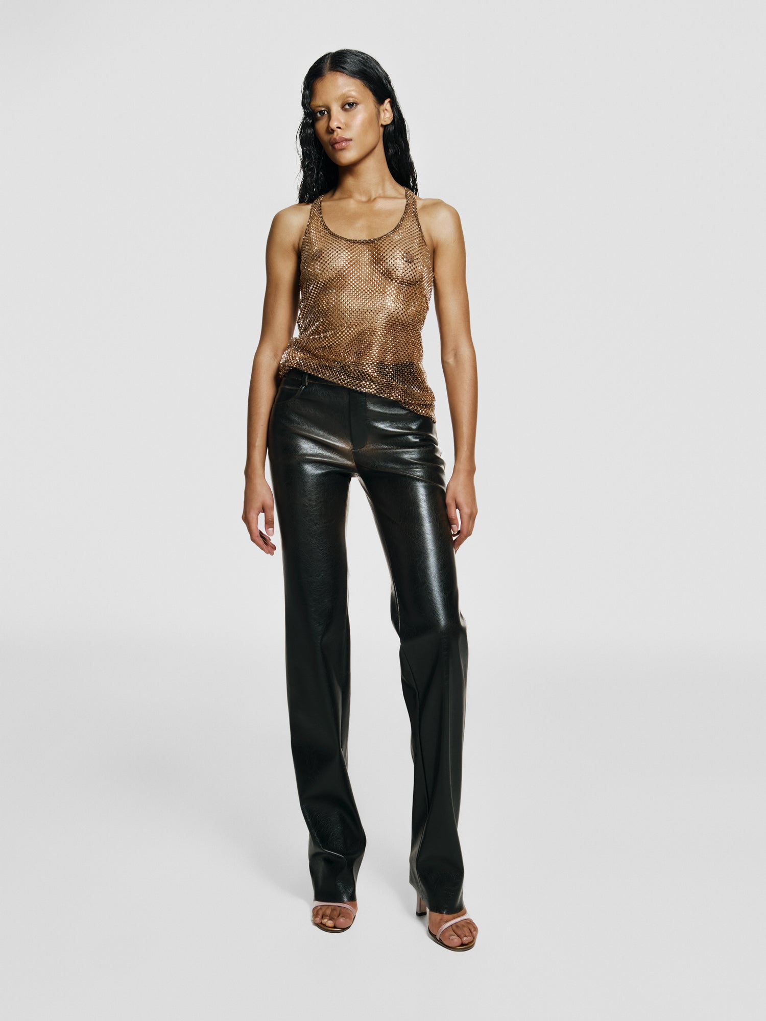 Full shot of a girl in a brown net tank top decorated with rhinestones and brown vegan leather medium rise straight leg pants