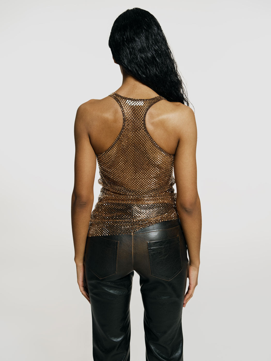 Medium full shot of a girl facing back in a brown net tank top decorated with rhinestones and brown vegan leather medium rise straight leg pants