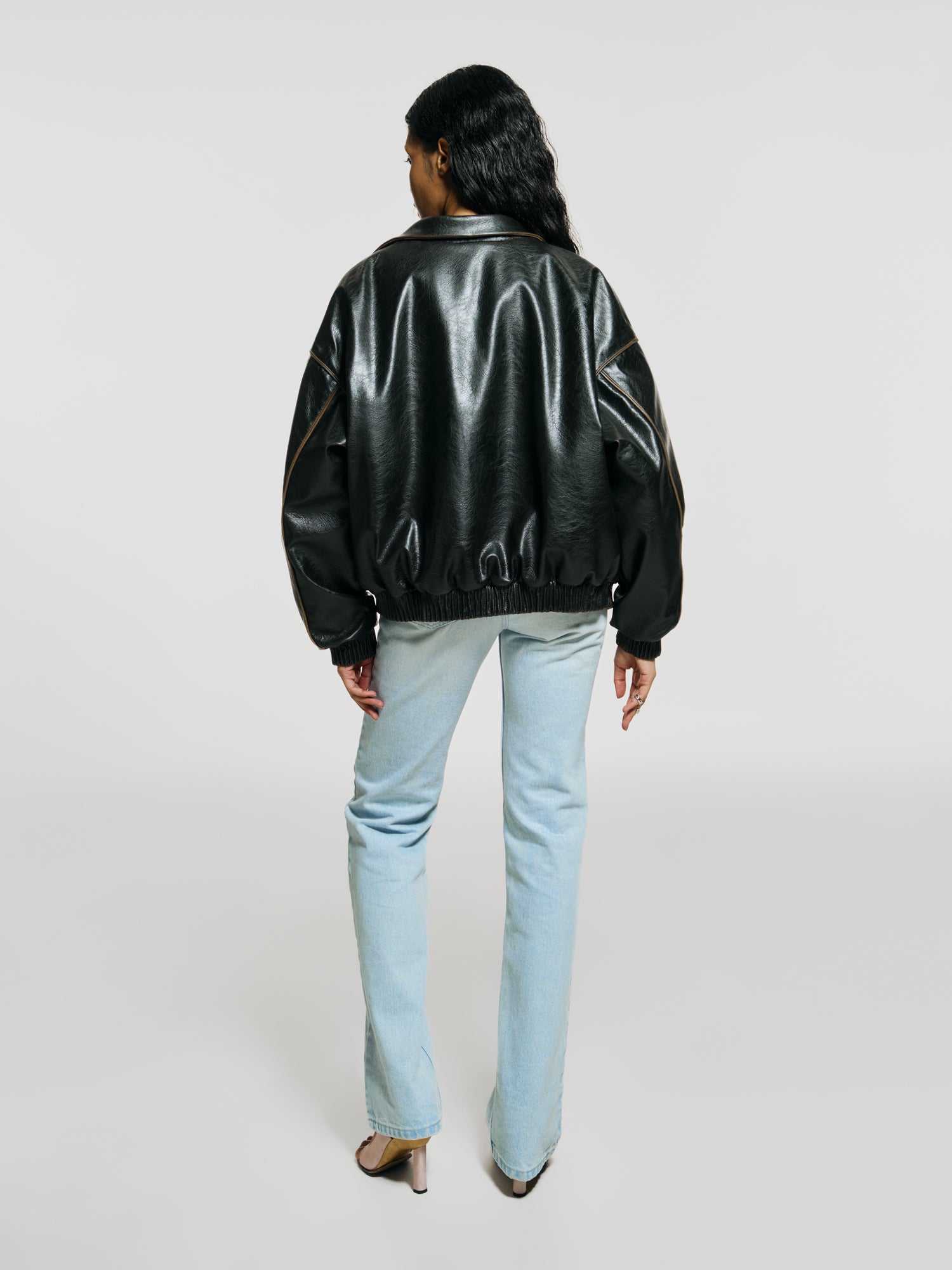 Full shot of a girl facing back in a brown vegan leather oversized bomber and blue high rise jeans with a straight leg