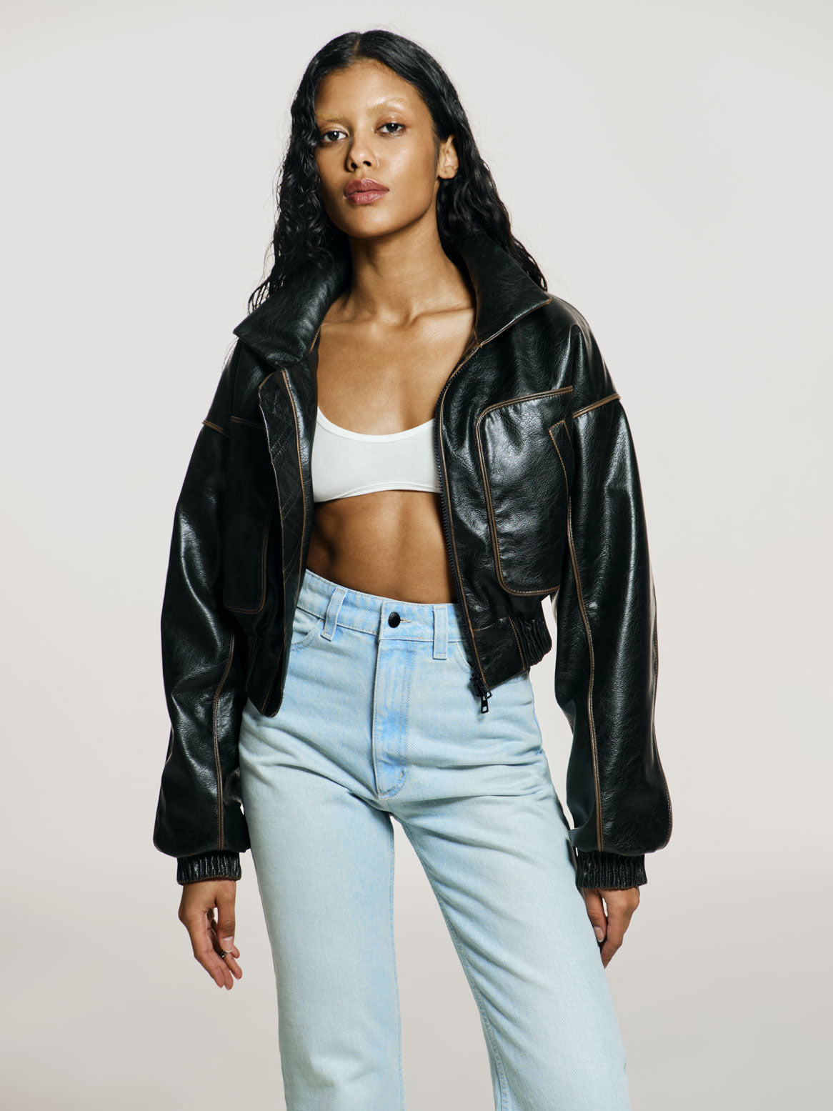 Medium full shot of a girl in a brown vegan leather oversized cropped bomber, white viscose crop top and blue high rise jeans with straight leg