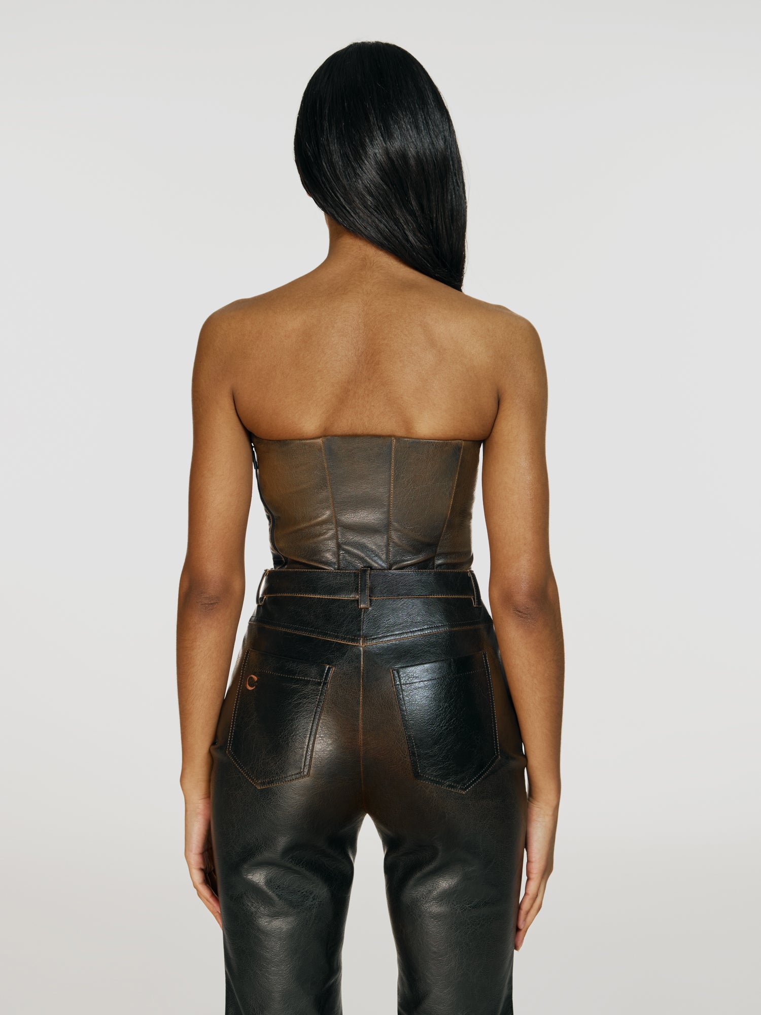 Cowboy shot of a girl facing back in a brown vegan leather tube top and brown vegan leather high rise pants with straight leg