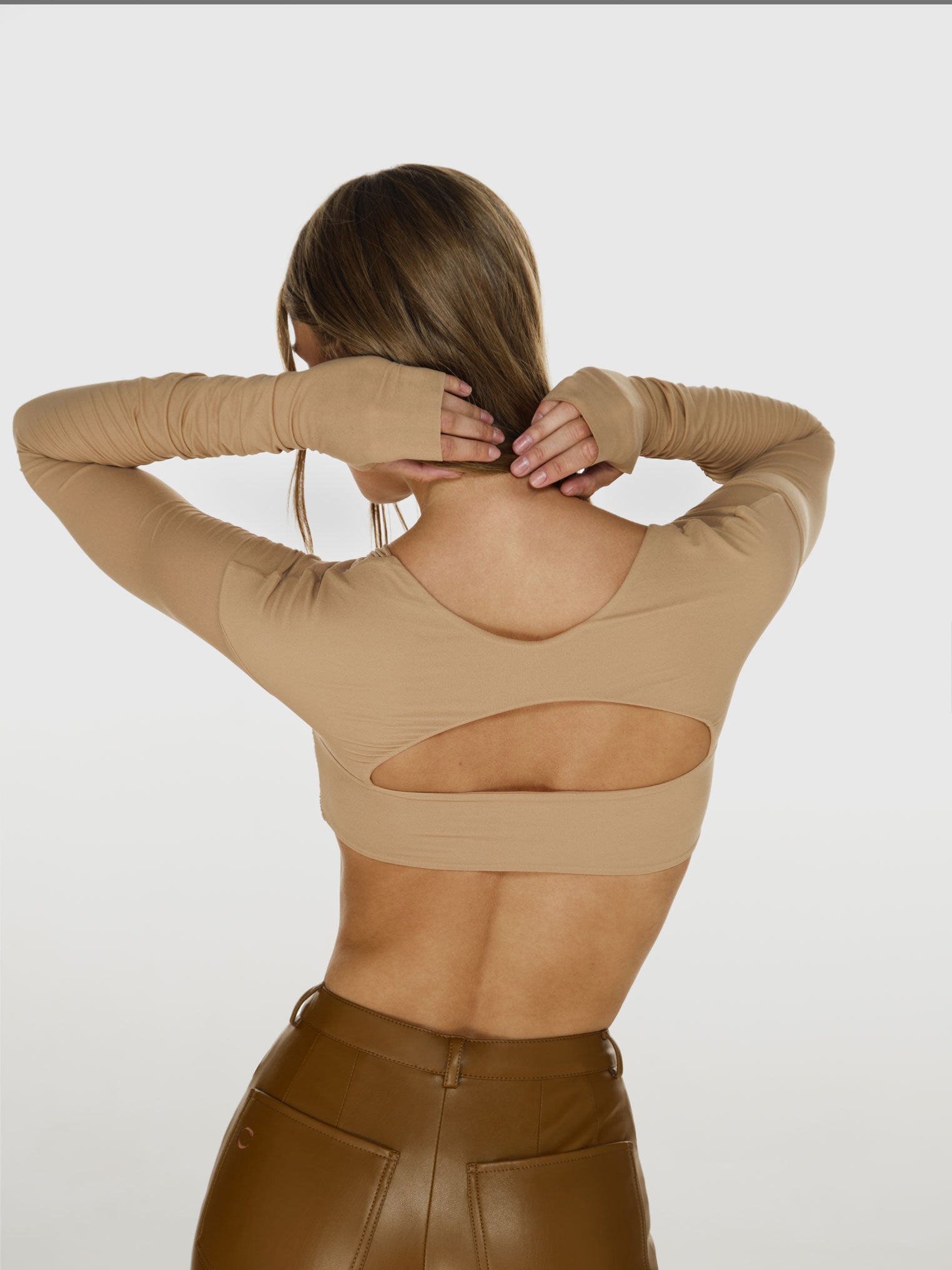 Medium shot of a girl in a beige long sleeved crop top, featuring horizontal cut at the back, and brown vegan leather pants with straight leg