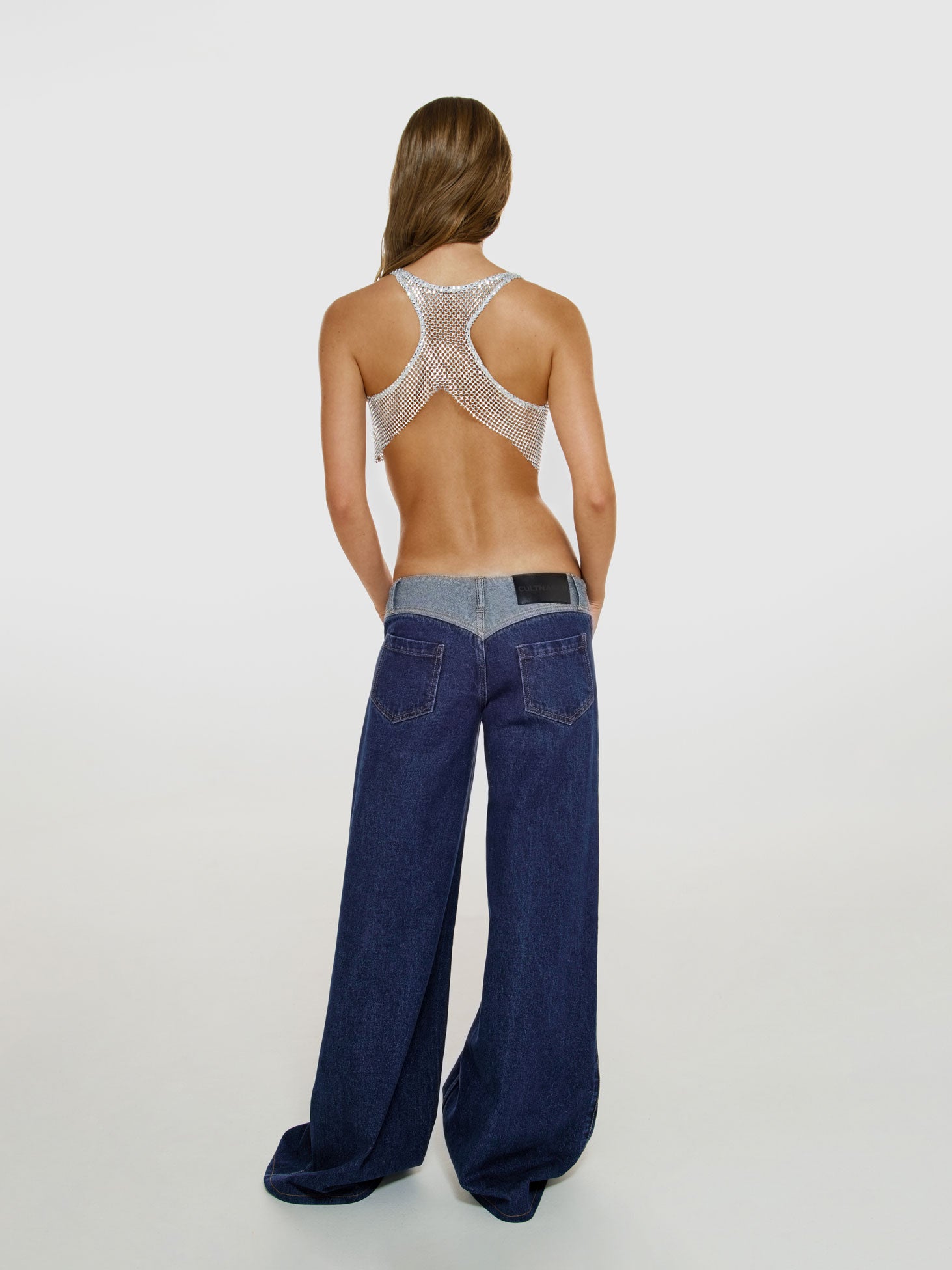 Full shot of a girl facing back in a grey net cropped tank top decorated with rhinestones and blue low rise wide leg jeans