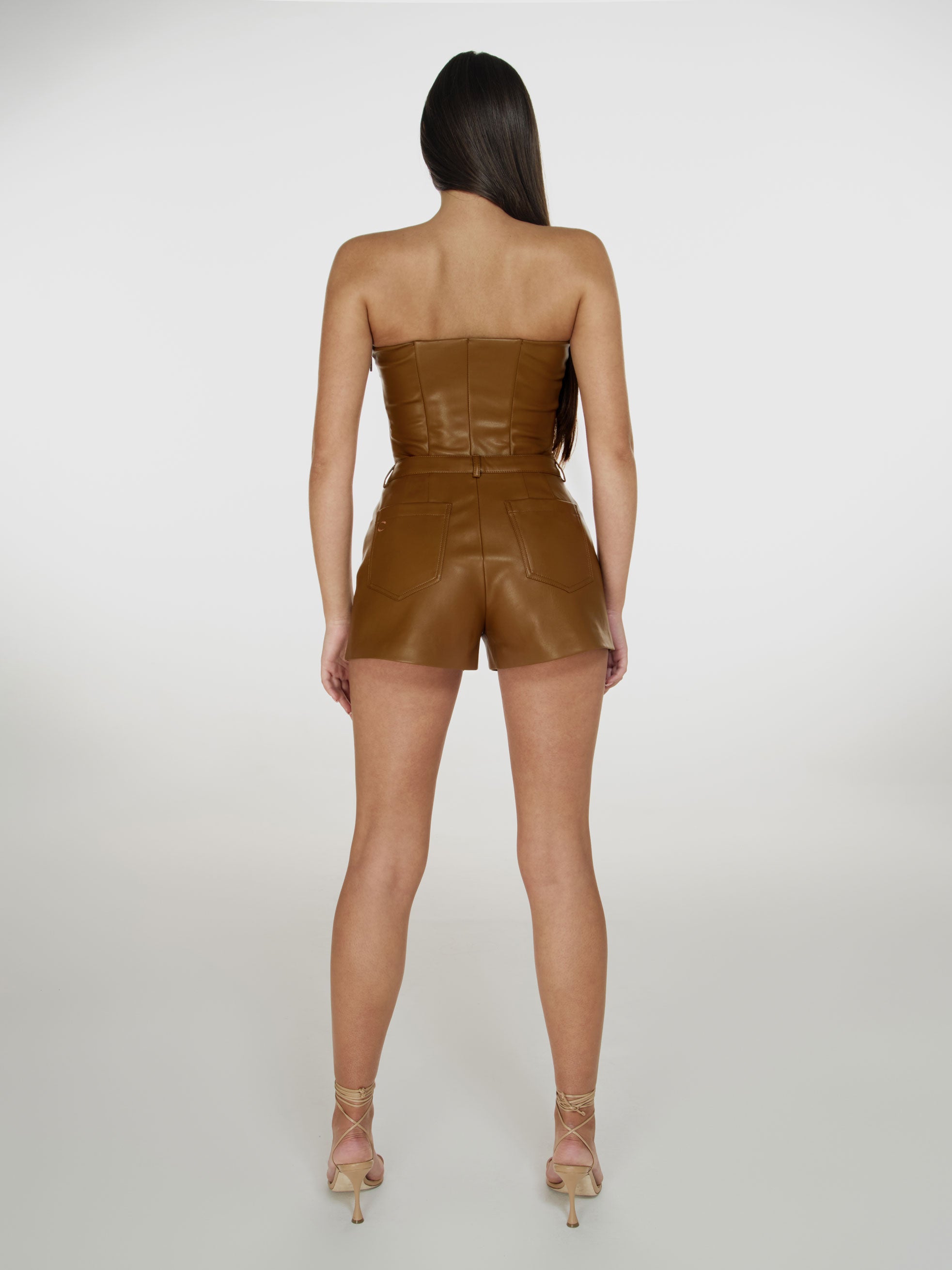 Full shot of a girl facing back in a brown vegan leather tube top and brown vegan leather high rise shorts