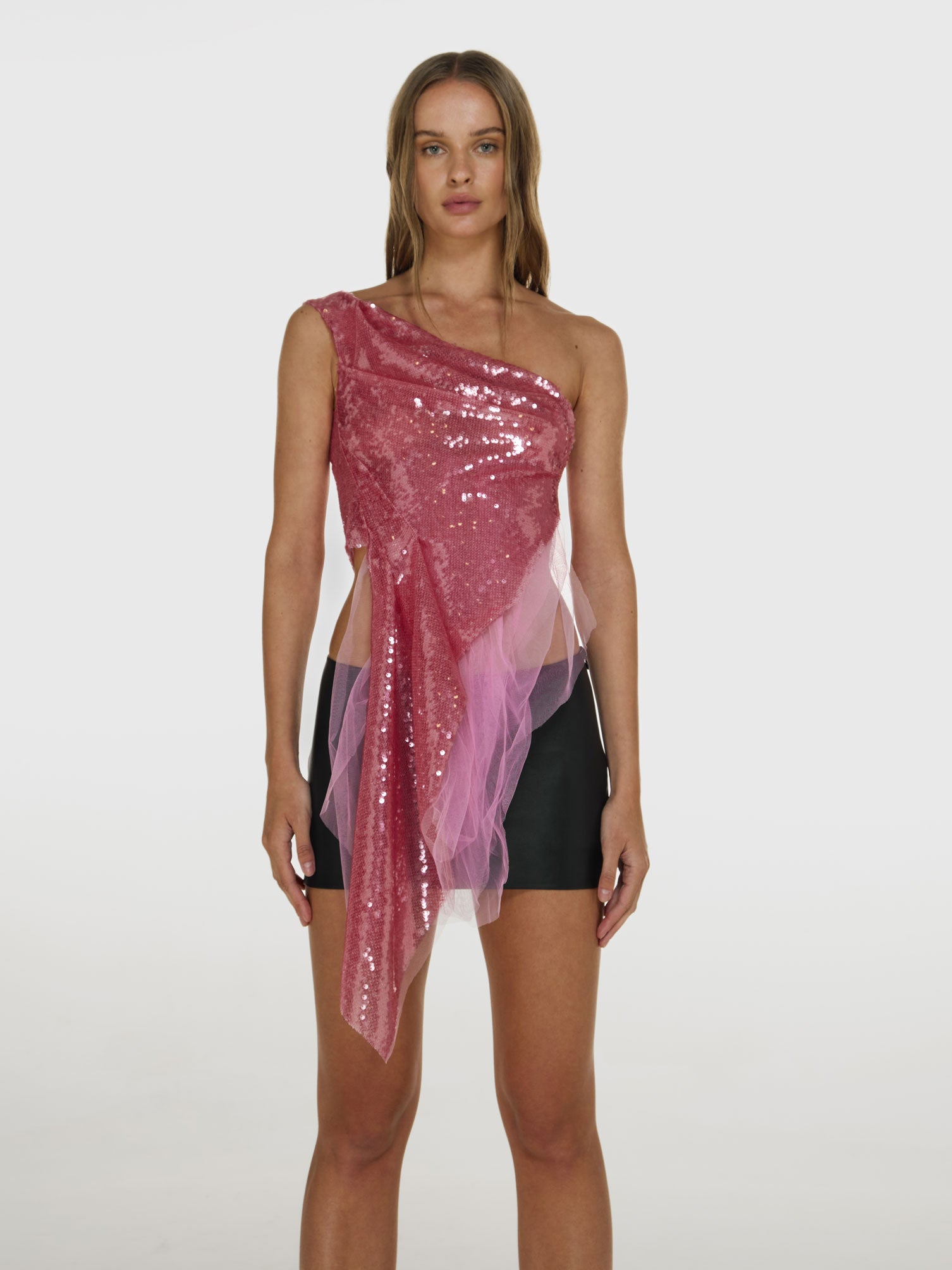 Medium full shot of a girl  in a pink sequinned one shoulder top with asymmetric hem in the front and a black vegan leather low rise mini skort