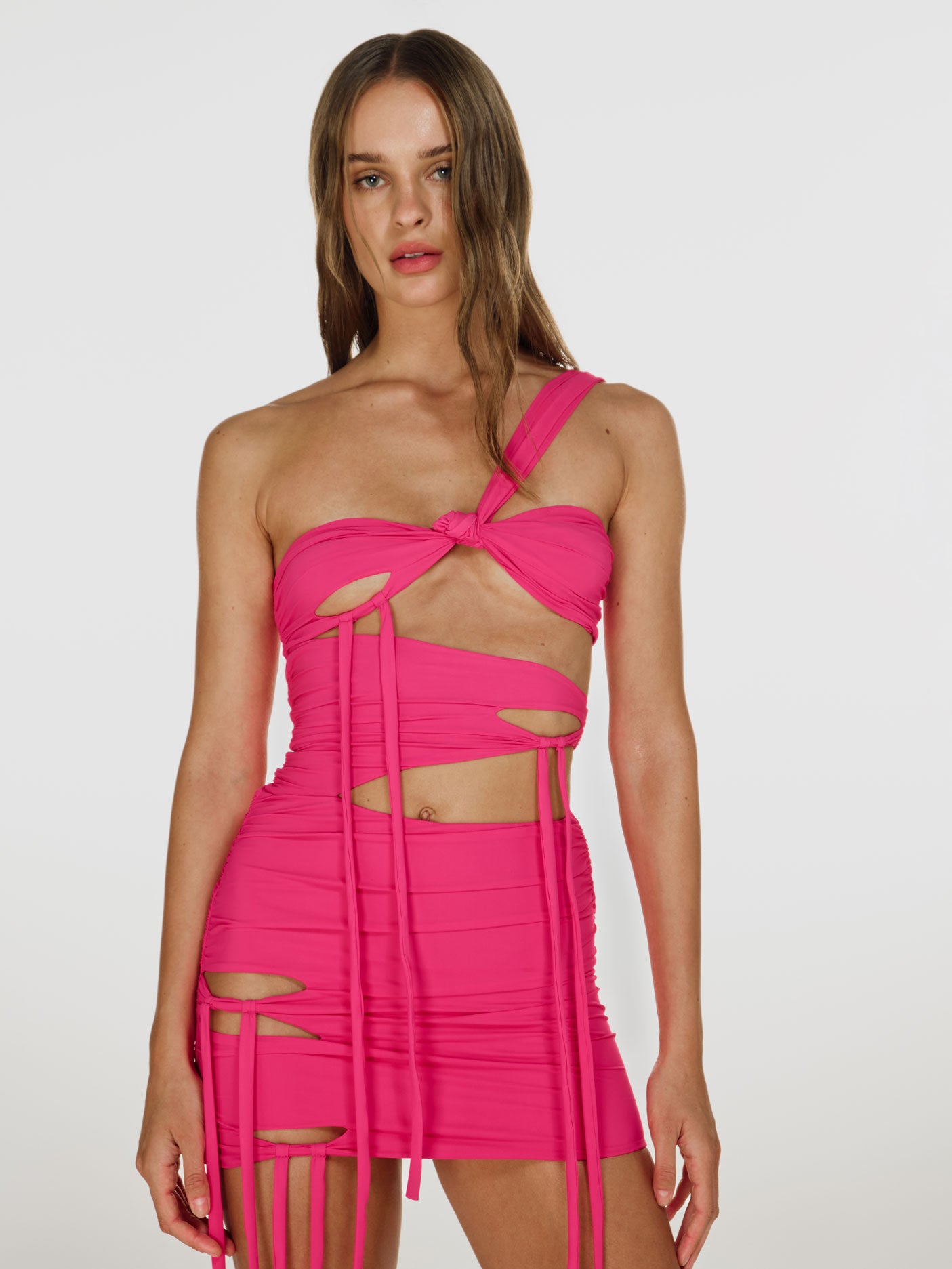 Cowboy shot of a girl in a pink one shoulder mini dress with extended laces, featuring horizontal cuts