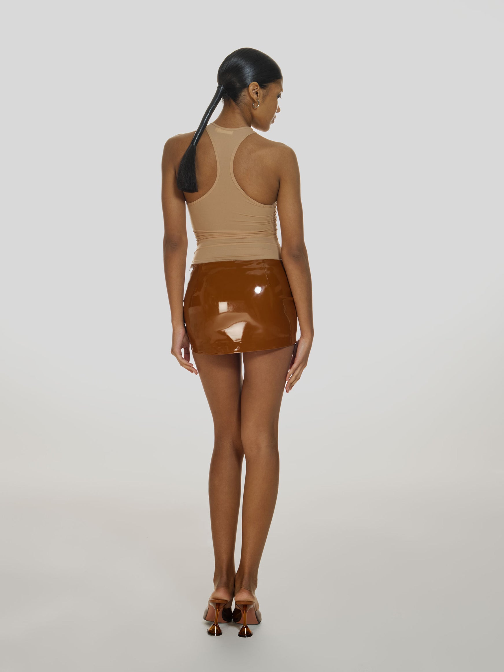 Full shot of a girl facing back in a brown vegan leather low rise mini skort, a beige sheer tank top and brown high heels