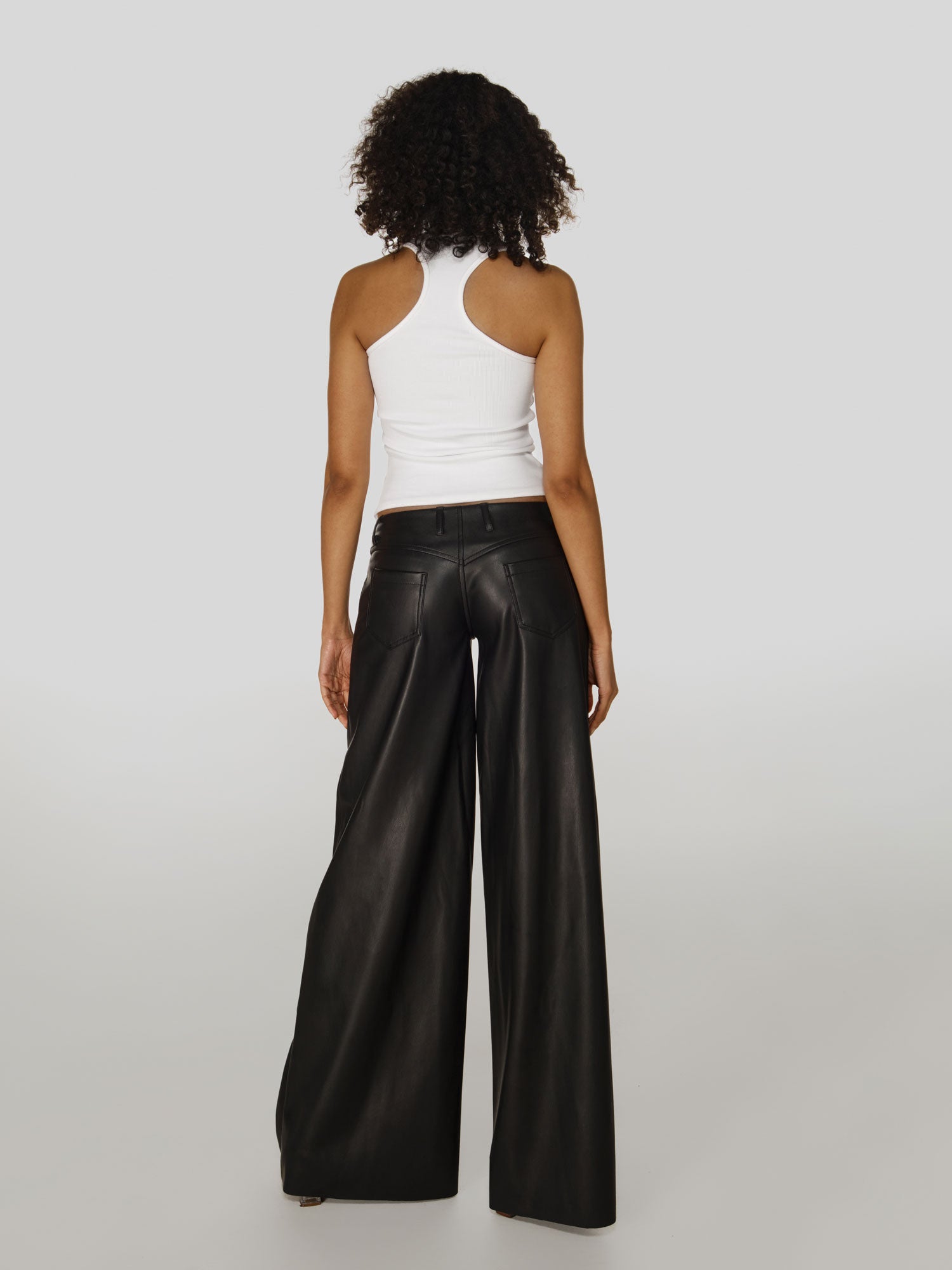 Full shot of a girl facing back  in a white viscose tank top and black vegan leather low rise wide leg pants