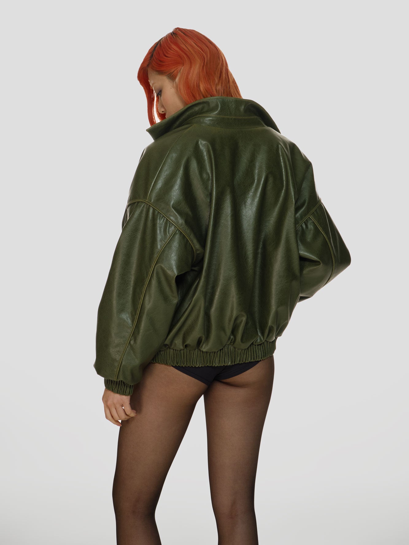 Medium full shot of a girl facing back in a green vegan leather oversized bomber, a white viscose crop top and black viscose high rise briefs
